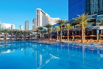 A large outdoor pool reflects the clear blue sky, surrounded by palm trees and lounge chairs. The backdrop showcases a modern resort structure with glass elements, providing a striking contrast against the serene water.