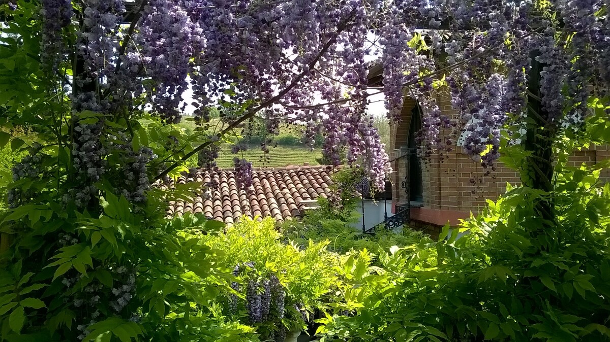 The image features a view framed by lush greenery and cascading wisteria flowers. A charming roof with terracotta tiles is visible in the background, leading to a traditional structure with an arched doorway. The vibrant colors of the garden enhance the serene countryside setting.