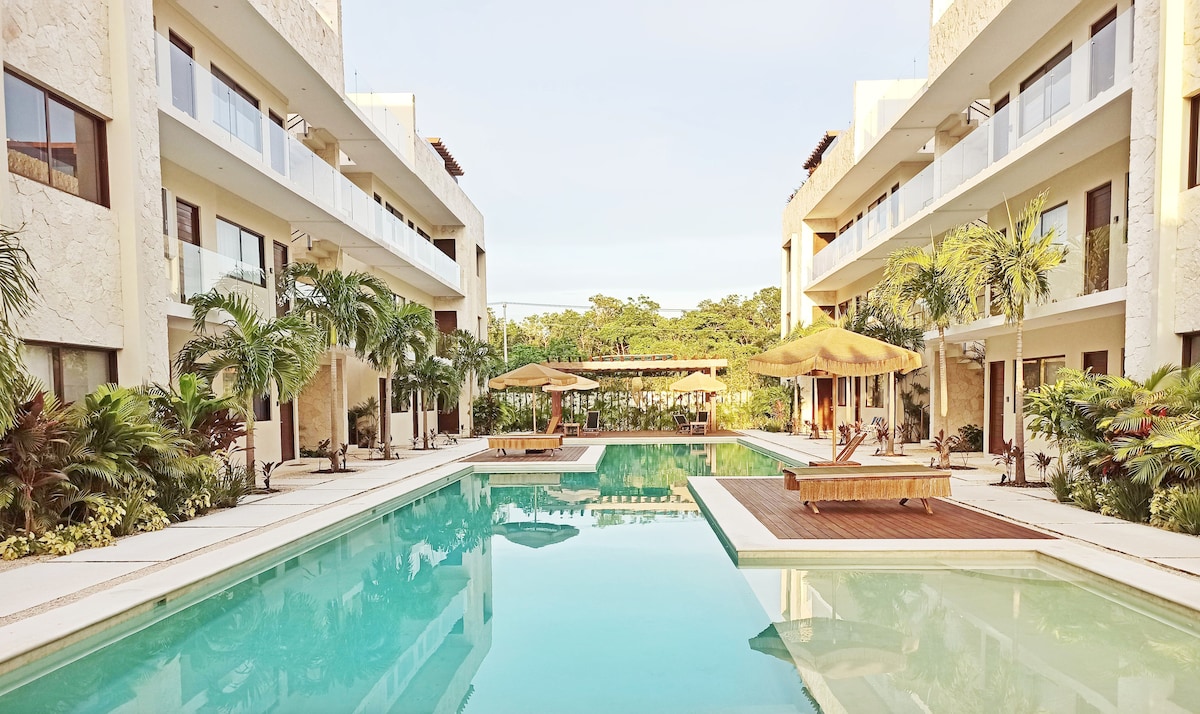 A spacious outdoor area features a long swimming pool surrounded by tropical greenery. Sun loungers and shaded seating areas are visible along the pool's edge, while two wooden platforms invite relaxation. The architecture showcases a modern design with balconies overlooking the inviting space.