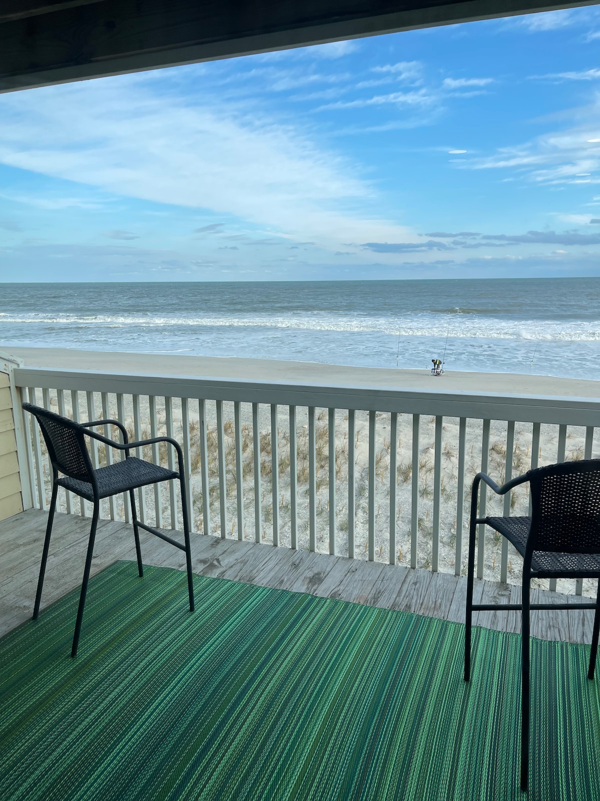 Two black chairs are positioned on a private deck, facing the ocean. The expansive view showcases the sandy beach, gently rolling waves, and a clear sky with scattered clouds. The deck features a green area rug, enhancing the inviting outdoor space.