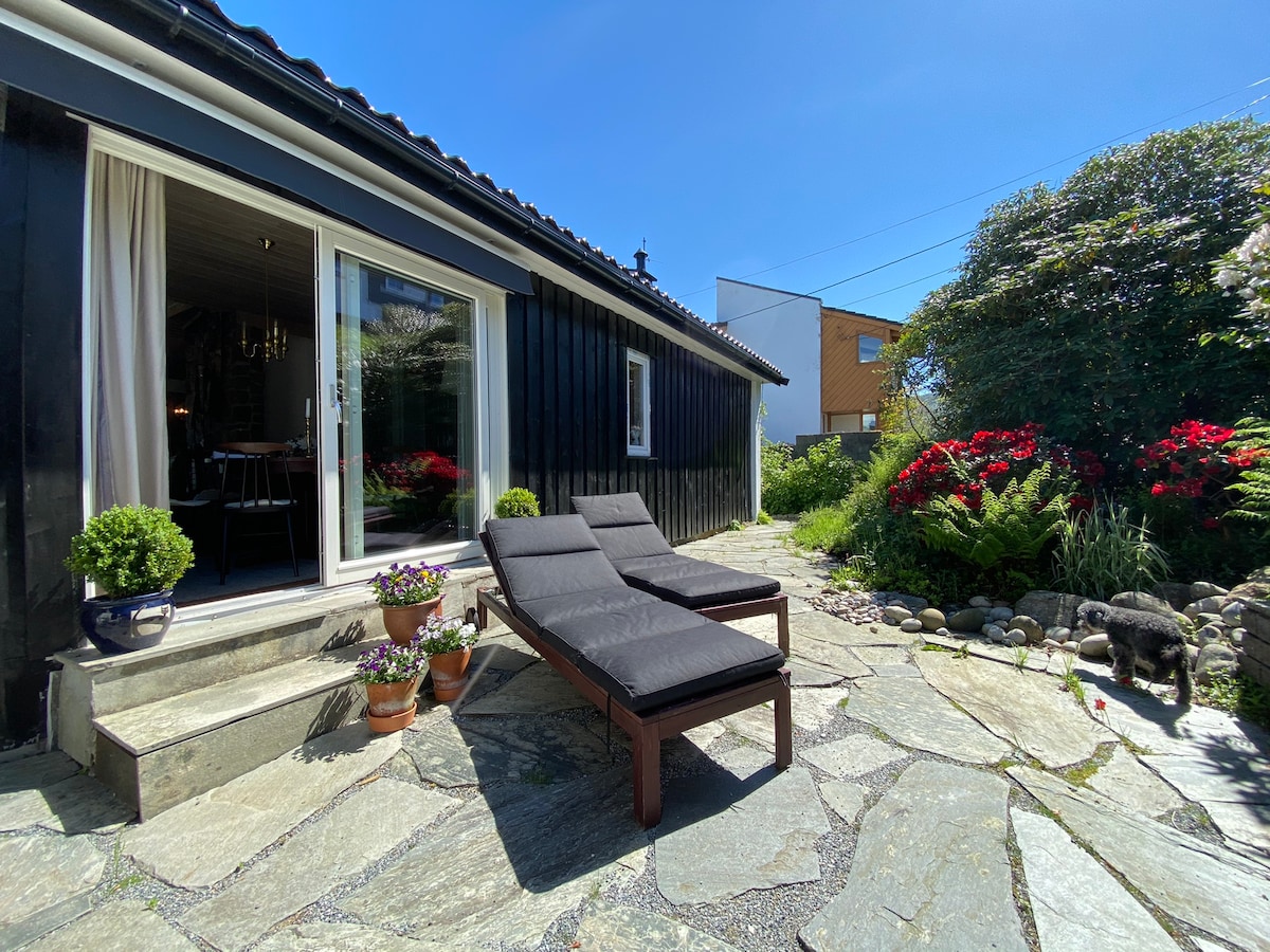 A stone patio area is shown with two dark lounge chairs arranged beside steps leading to a black wooden house. Potted plants display colorful blossoms, while a neatly landscaped garden with vibrant greenery and flowers creates a serene outdoor setting.