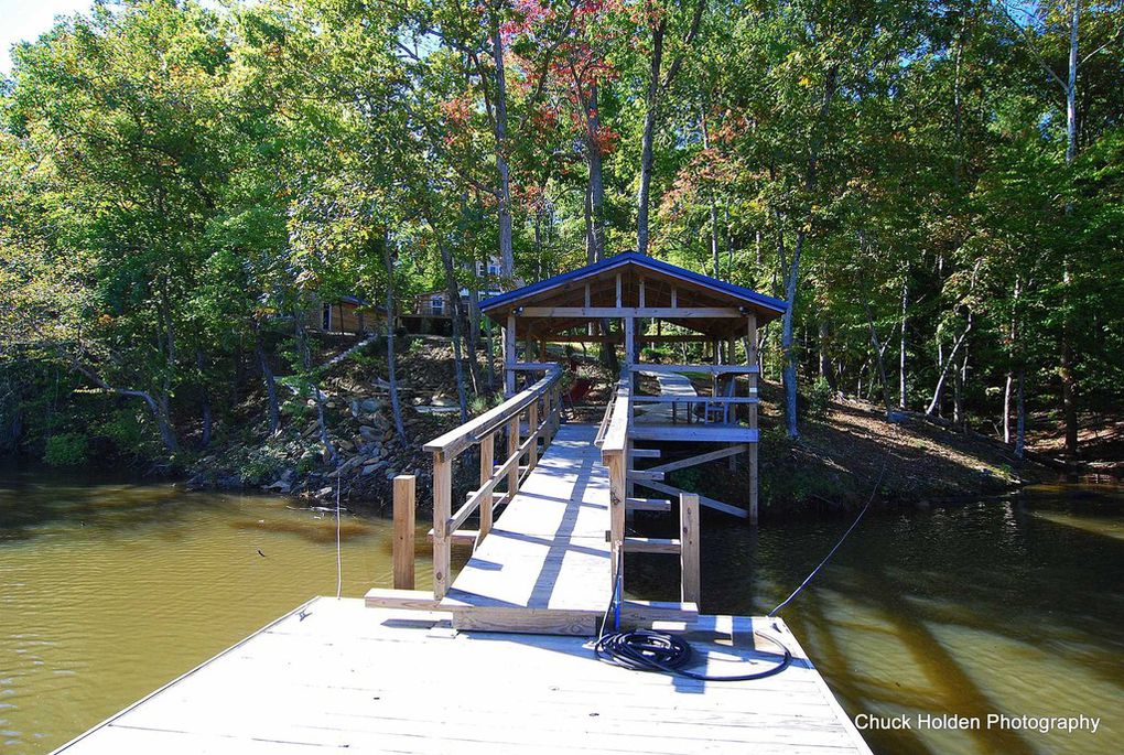 A wooden dock extends over the water, leading to a covered boat slip. Lush green trees frame the area, providing shade. The calm lake reflects the surrounding landscape, inviting opportunities for fishing and water sports.