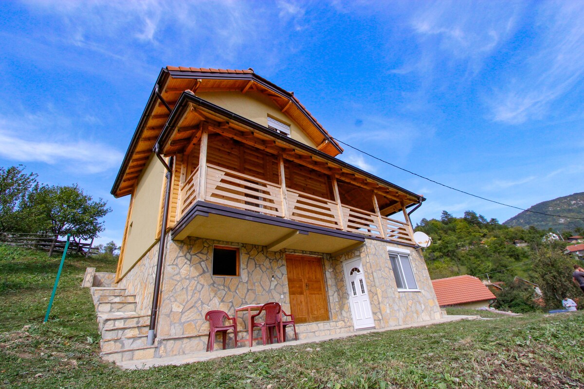 A two-story house features a stone exterior with a wooden balcony overlooking the surrounding landscape. Two red chairs are positioned on the patio, and a well-defined entrance showcases a wooden door framed by white accents.