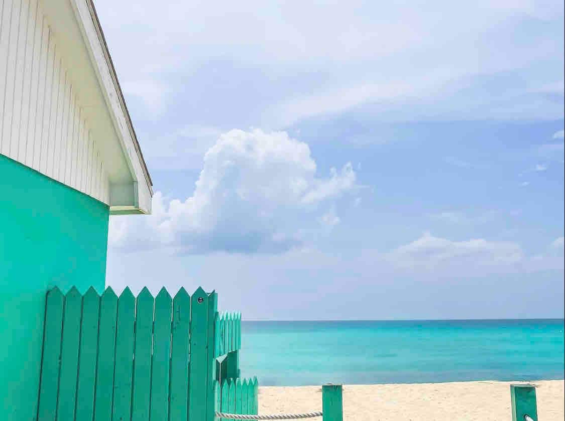 A vibrant turquoise fence surrounds a beach access point, leading to golden sand that meets the calm turquoise sea. A few fluffy clouds drift in the sky, enhancing the serene coastal ambiance.