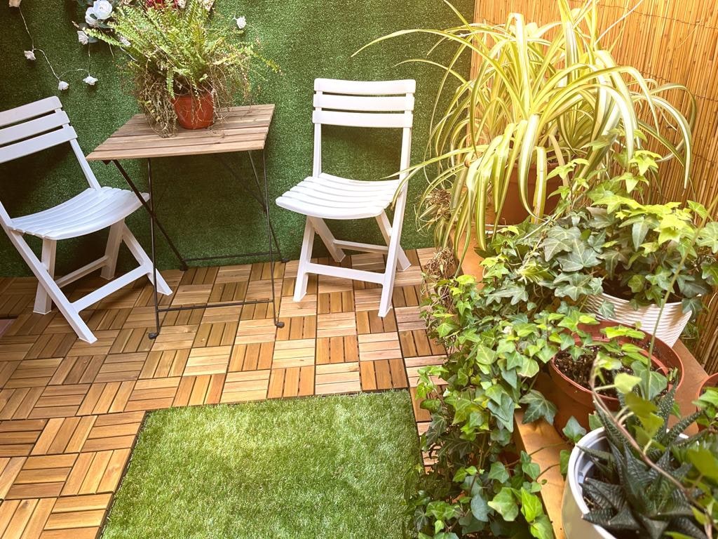 A private patio is presented with green walls and wooden flooring. Two white chairs are arranged around a small table, surrounded by various potted plants and greenery. A patch of artificial grass adds a touch of color, enhancing the tranquil outdoor space.