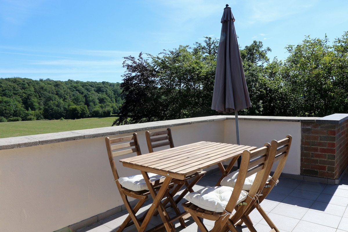 An outdoor terrace is featured, equipped with a wooden table and four matching chairs. A large umbrella provides shade, while scenic views of lush greenery stretch out in the background under a clear blue sky.