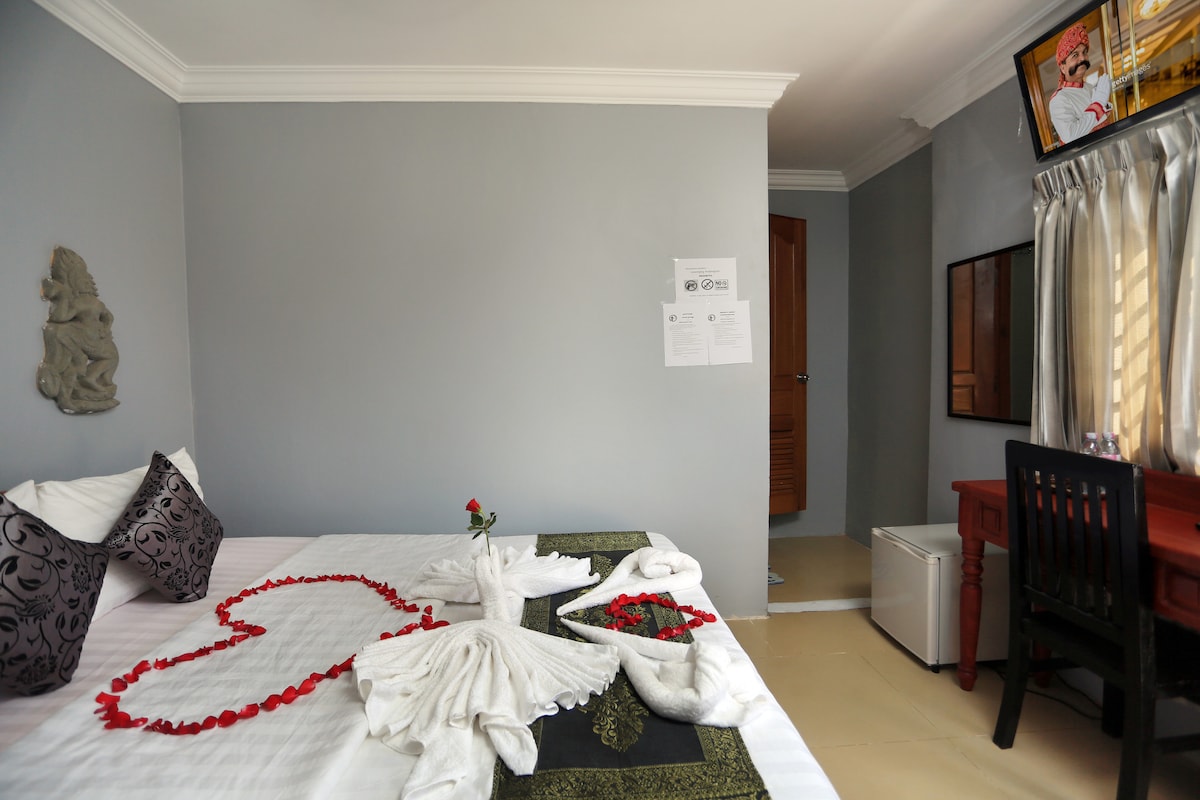 A cozy room features a spacious bed with decorative towel arrangements in the shape of a heart, surrounded by fresh rose petals. A small table and chair provide a work area, while a mini fridge is visible. Natural light enters through sheer curtains.