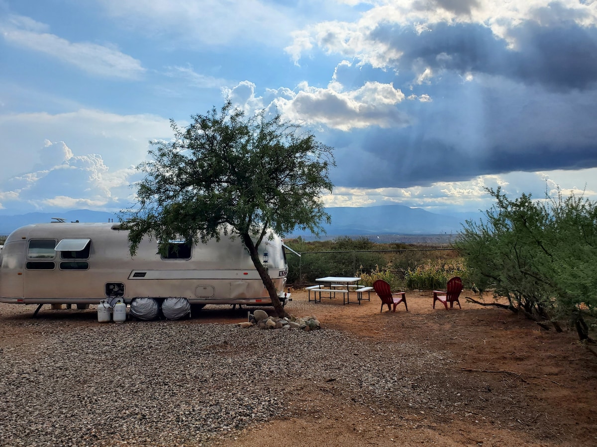 A vintage Airstream is situated on a gravel plot surrounded by desert vegetation. Two red chairs are positioned in front of the trailer, with a picnic table nearby. A scenic view of distant mountains is visible under a partially cloudy sky.