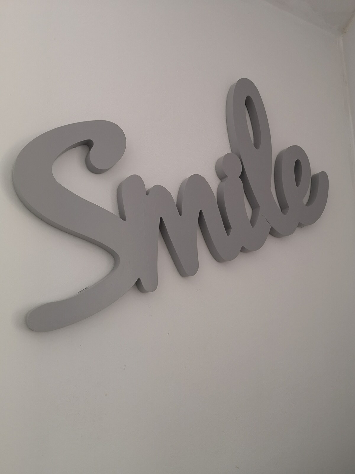 A grey wall decoration features the word 'Smile' in a playful, cursive design. The piece is mounted on a simple white wall, creating a light and inviting focal point within the space.