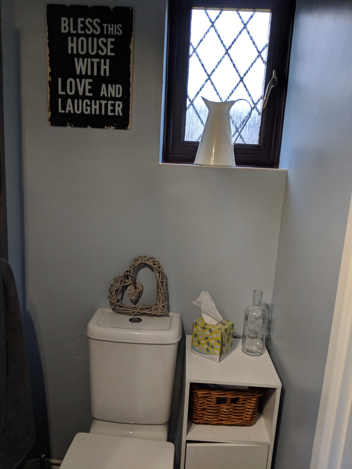 A neatly arranged bathroom corner is depicted, featuring a white toilet with a decorative heart-shaped accent made of wicker placed on top. A small wicker basket and a glass bottle are positioned on a white cabinet, alongside a tissue box and a vase on the windowsill.