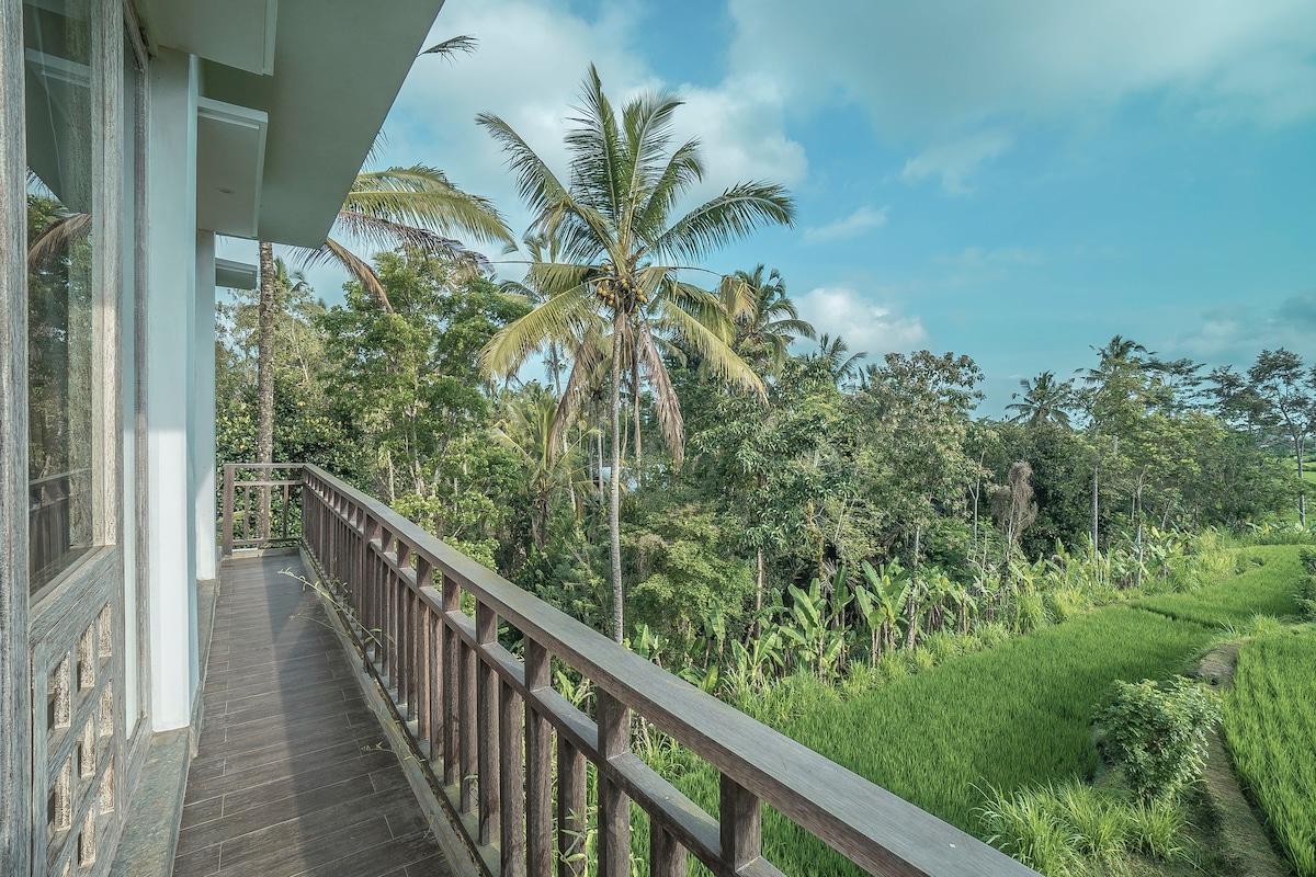 A spacious balcony extends from the villa, providing unobstructed views of lush greenery and rice fields. Tall palm trees sway gently in the breeze, contributing to the serene atmosphere of the surrounding tropical landscape.