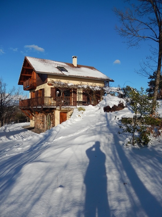A chalet surrounded by snow-covered landscapes is depicted, showcasing a two-story structure with wooden accents. The exterior features a balcony and a stone base, contrasting against the white snow. Clear blue skies are visible in the background, highlighting the serene winter setting.