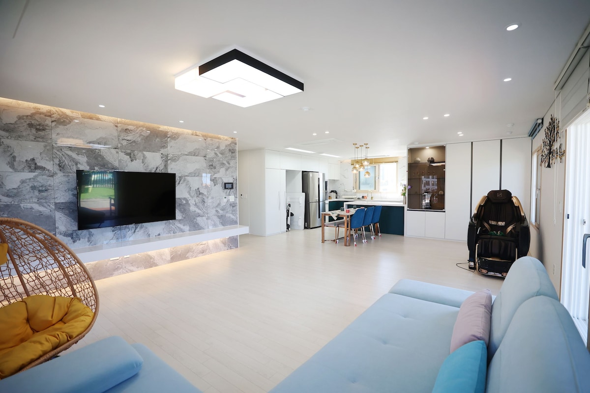 A spacious living area is highlighted by a large television mounted against a stone accent wall. Light wood flooring complements the modern furnishings, including a soft blue sofa. A massage chair is positioned nearby, and a contemporary kitchen area is visible in the background.