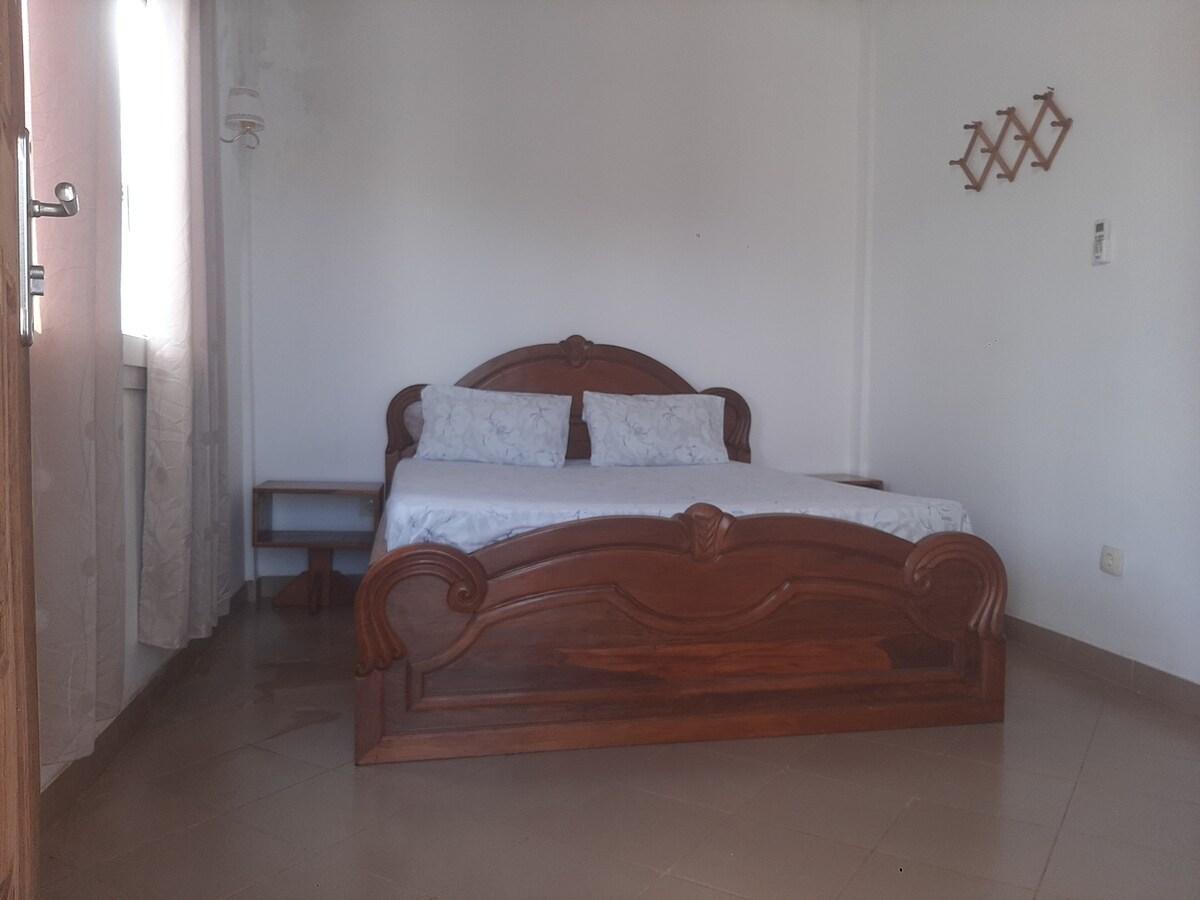 A spacious bedroom featuring a wooden bed with a decorative headboard and two pillows arranged neatly. A small bedside table is placed next to the bed, and natural light enters through a window adorned with sheer curtains. Wall hangers are visible for additional storage.