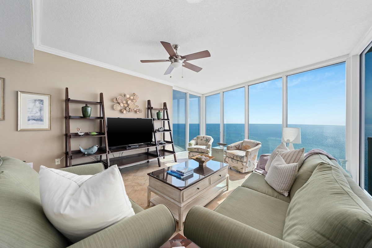 A spacious living area features a large sectional sofa and a decorative coffee table. Two armchairs are positioned to take in views of the Gulf, framed by floor-to-ceiling windows that allow natural light to fill the space.