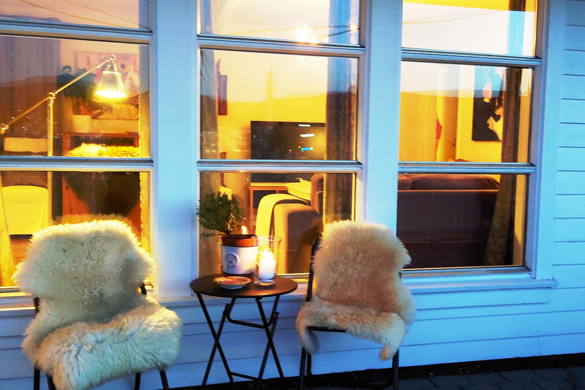 A cozy outdoor seating area is presented with two furry chairs positioned beside a small table. A candle is placed on the table, creating a gentle glow, while the inviting interior of the apartment is visible through large windows.