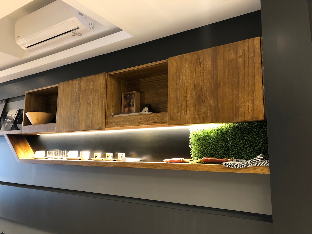 A wooden shelf is mounted on a grey wall, illuminated by soft lighting. It hosts decorative items, including a bowl and small photographs, alongside lush green accents. The arrangement creates a functional yet appealing space, suitable for both relaxation and social interaction.