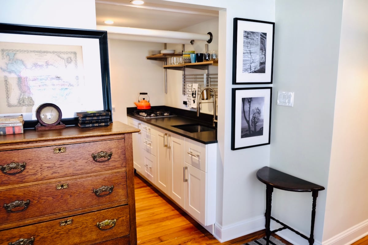 A compact kitchen area is highlighted by sleek cabinetry and modern appliances. Light-colored walls are complemented by dark countertops. A wooden dresser and a small side table enhance the space, while black-and-white artwork is displayed on the adjacent wall.