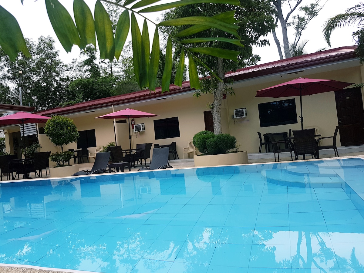 A refreshing pool area features clear blue water, surrounded by seating options that include lounge chairs and shaded tables with umbrellas. Lush greenery and trees create a natural backdrop to the inviting space, enhancing the overall relaxing ambiance.
