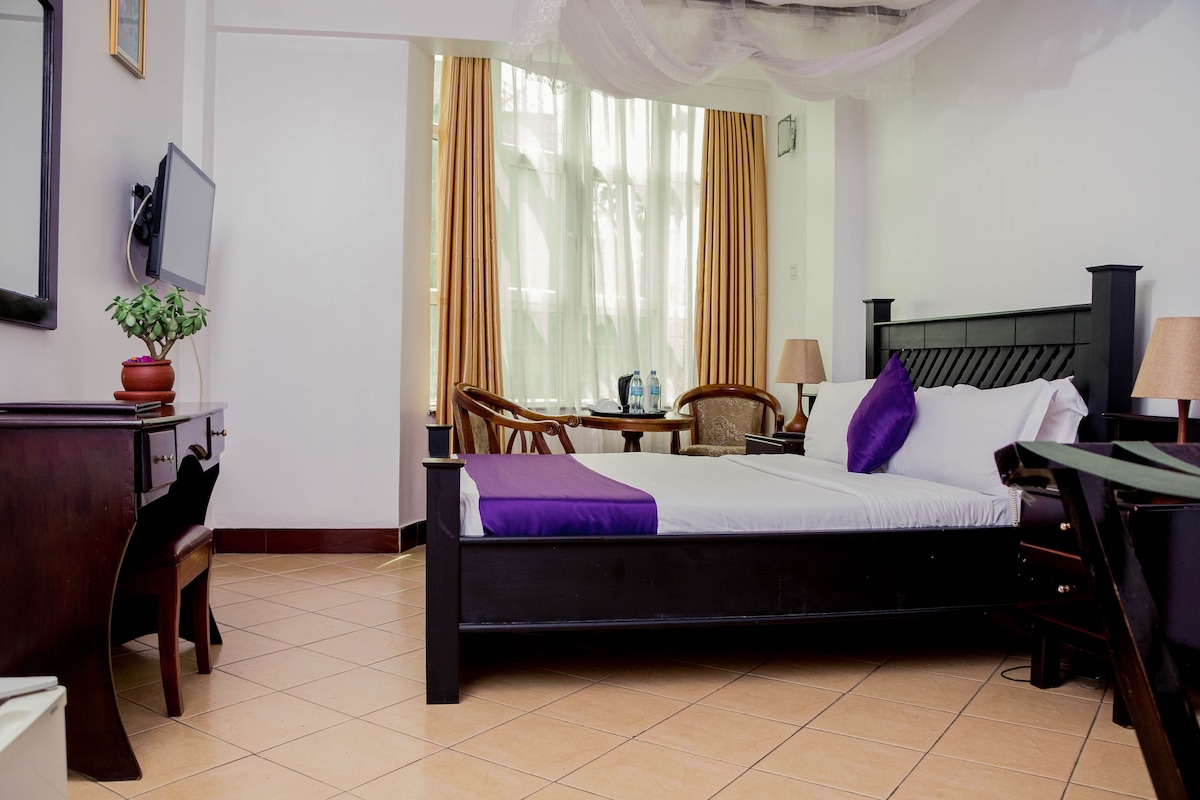 A single room featuring a spacious bed with white linens and a purple accent pillow. Natural light streams in through large windows adorned with light-colored curtains. A wooden desk and chair, along with a small dining table and chairs, are positioned thoughtfully in the room.