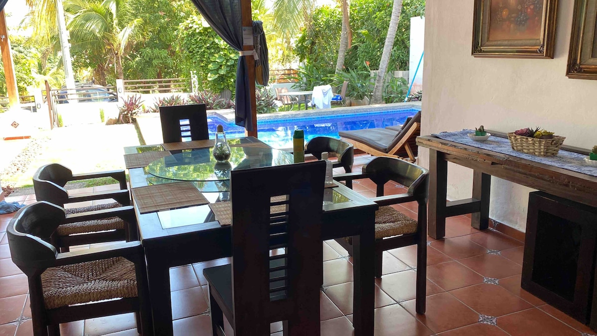 An outdoor dining area is presented, featuring a dark wooden table surrounded by six black chairs. A stone pathway leads to a swimming pool framed by lush greenery, creating a serene environment. Natural light filters through the surrounding foliage, enhancing the tranquil setting.