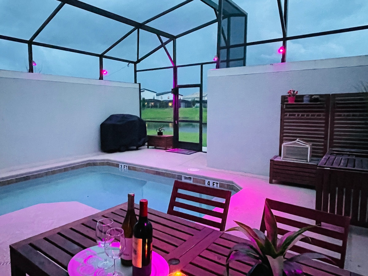 A private mini heated pool is surrounded by a screened enclosure, featuring LED lighting in purple tones. A dining area is set with a table and chairs nearby, while a barbecue grill is positioned to the side. The tranquil lake view is visible through the enclosure.