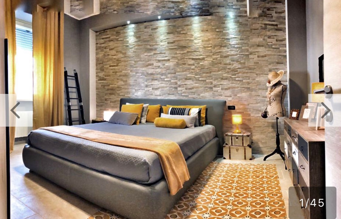 A spacious bedroom features a textured stone accent wall, with soft lighting enhancing its calm atmosphere. A large bed is dressed with decorative pillows and a warm throw blanket. Textured tiles cover the floor, complementing the modern furnishings, including a bedside table and a stylish clothing display.