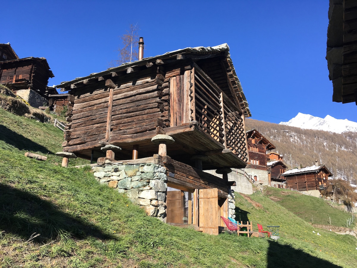 An alpine structure, elevated on stone pillars, features a traditional wooden design with a sloping roof. The surrounding landscape showcases lush green grass and scattered wooden chalets, with a backdrop of snow-capped mountains under a clear blue sky.