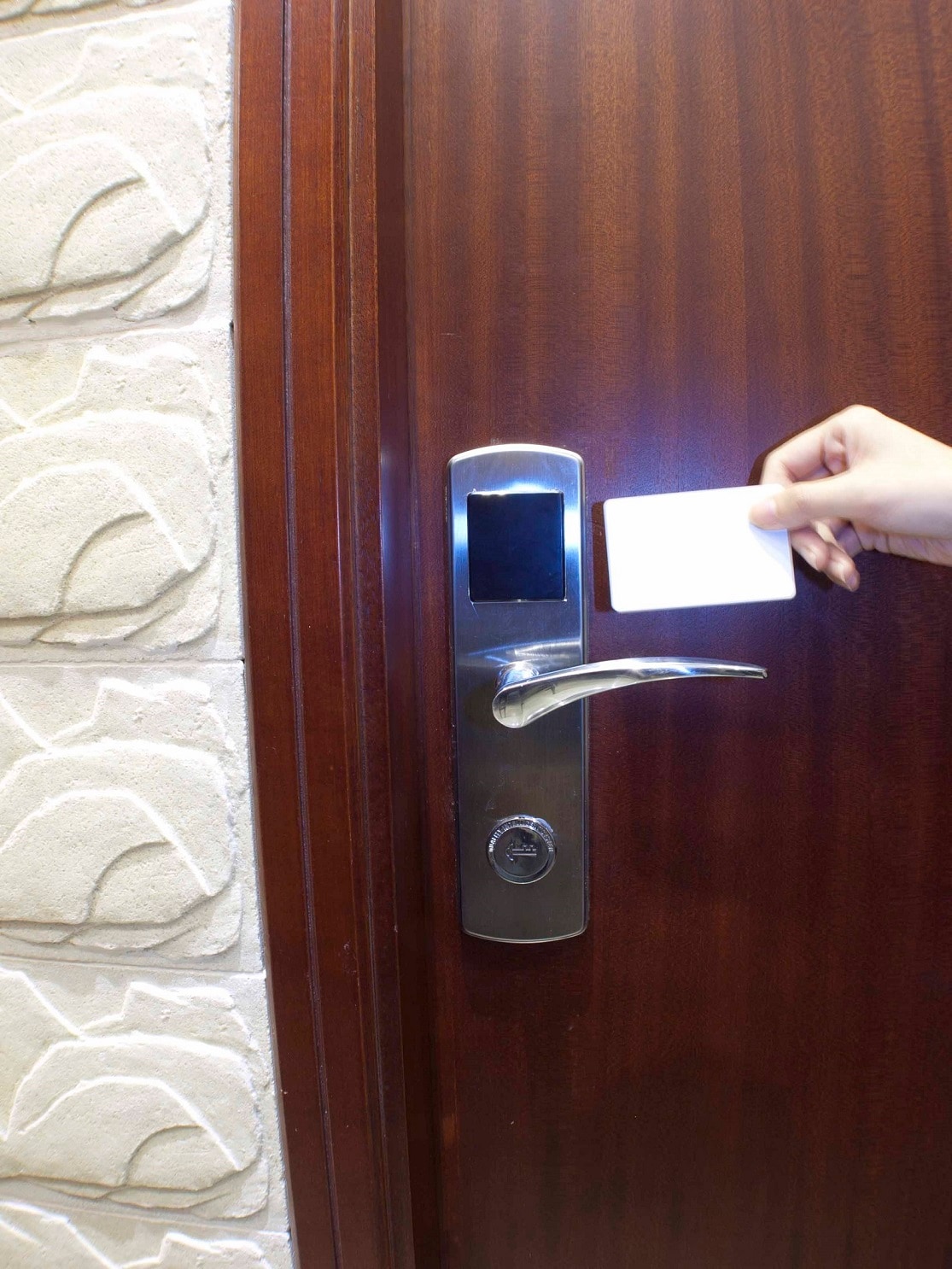 A wooden door with a sleek metallic handle and digital lock is shown, as a hand holds a key card near the sensor. A textured stone wall is visible beside the door, providing a contrast to the smooth wood.