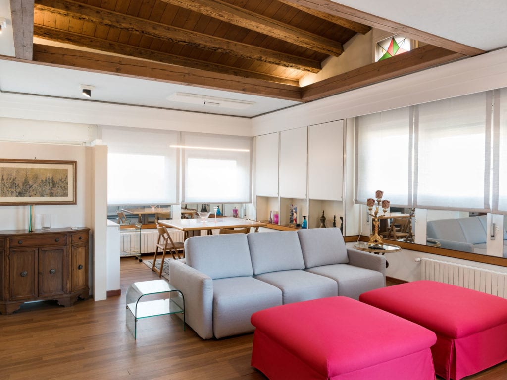 A spacious living area features wooden ceiling beams and large windows that allow ample natural light. A gray sofa is paired with two vibrant pink ottomans. A dining table is set in the background, complemented by decorative accents and a wooden sideboard.
