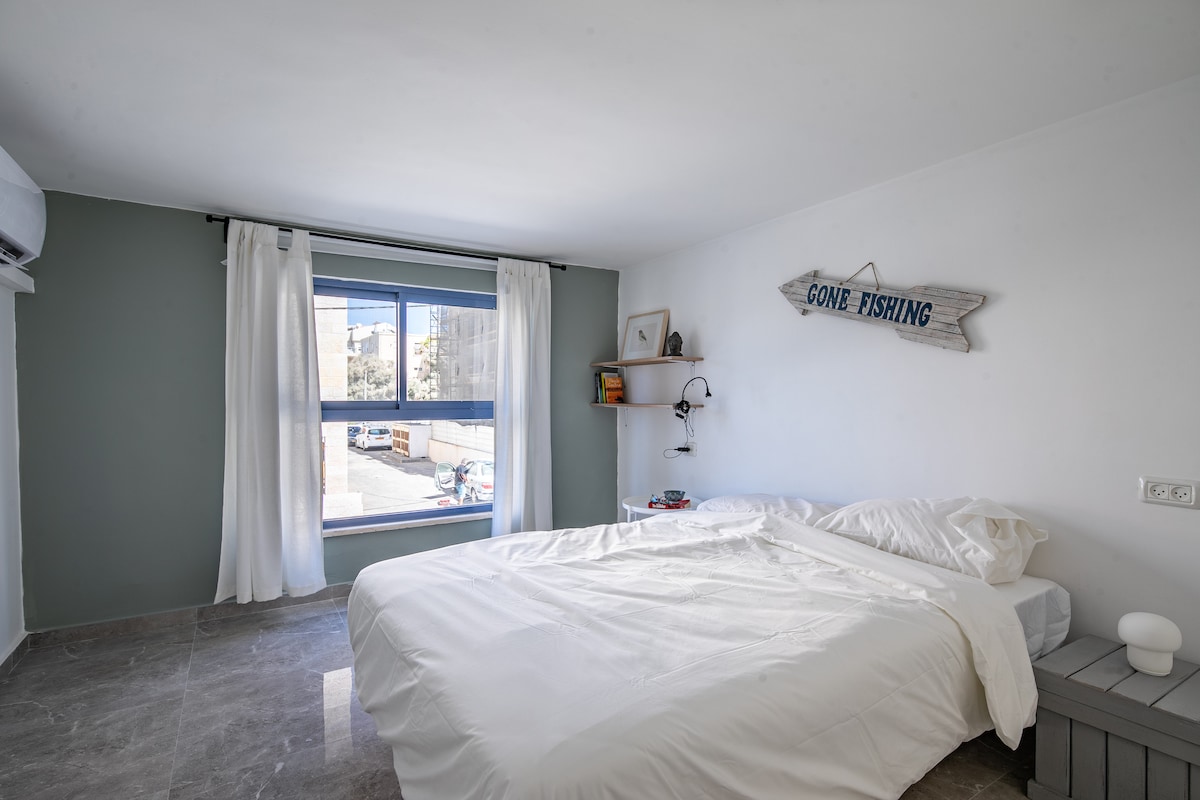 A well-lit bedroom features a spacious bed dressed in crisp white linens. A 'Gone Fishing' sign adds character to the softly colored wall. Natural light enters through a large window, framed by sheer curtains, while a bedside lamp and small shelf provide functional decor.