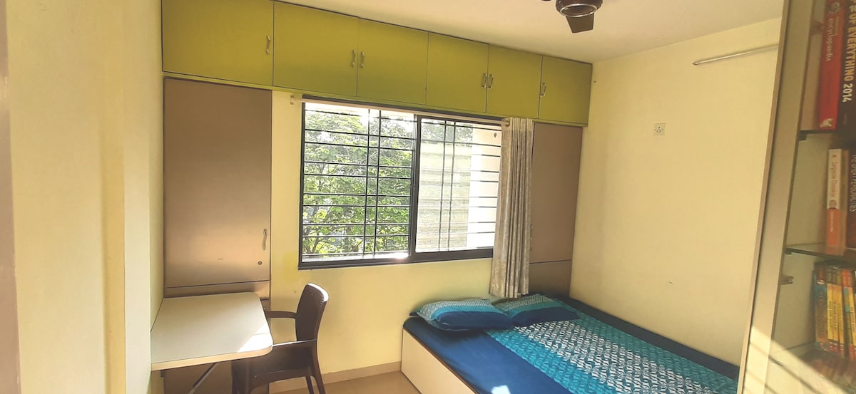 A well-lit bedroom features a spacious bed with a blue coverlet and a window with a view of greenery. A desk and chair provide a functional workspace, while built-in cabinets offer ample storage. A ceiling fan adds comfort to the air quality in the room.