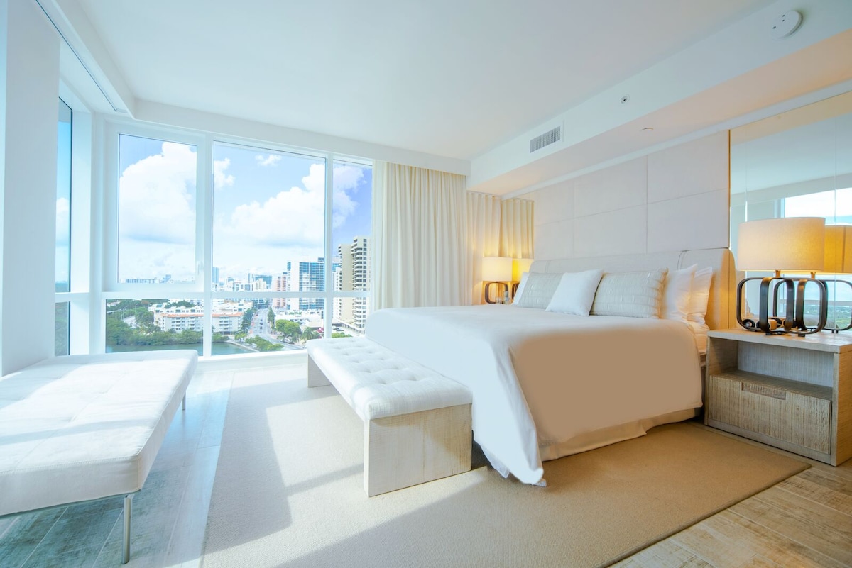 A spacious bedroom is highlighted by a plush king-sized bed and two contemporary lamps on bedside tables. A large window offers a panoramic view of the city skyline and natural light fills the room, complemented by soft neutral tones and textures.