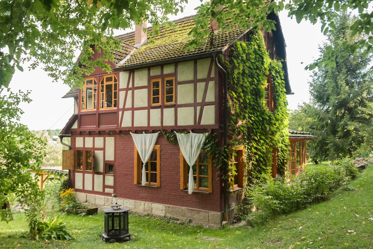 A charming, restored house displays a combination of half-timbered and brick architecture, surrounded by lush greenery. Large windows are adorned with light curtains, and a small lantern is positioned on the grass in front, enhancing the tranquil outdoor space.