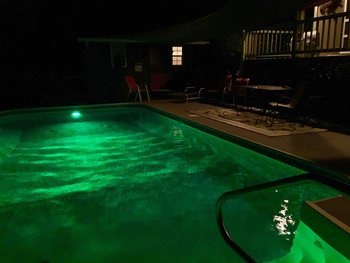 A well-lit pool is shown at night, featuring an inviting turquoise glow. Lounge chairs and a dining set are positioned nearby on a surface covered with outdoor decor. Soft light is visible emanating from the house in the background, providing a sense of warmth.