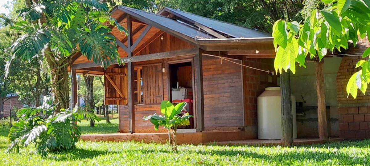 A rustic cabin is set within a lush green landscape, featuring wooden beams and a partially open design. Tropical plants surround the space, and a large water tank is visible beside the structure. The natural environment enhances the serene atmosphere.