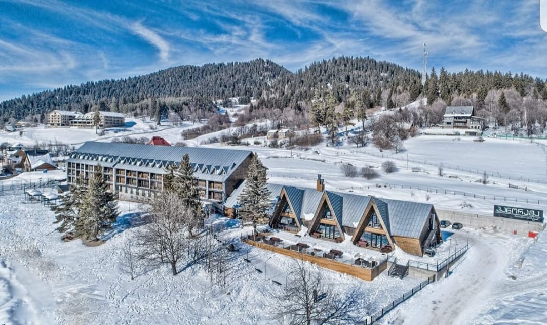 An aerial view of the Hotel showcases a modern structure surrounded by a blanket of snow. Snow-covered trees and rolling hills create a serene backdrop. Two distinctive roofs can be seen, complementing the natural landscape, while clear blue skies provide a bright atmosphere.