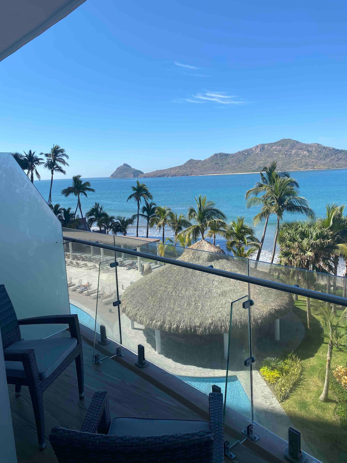 A balcony offers a stunning view of the blue ocean and distant mountains, framed by tall palm trees. Comfortable seating faces the expansive shoreline, while a thatched-roof structure is partially visible below. The scene captures the tranquil coastal atmosphere.