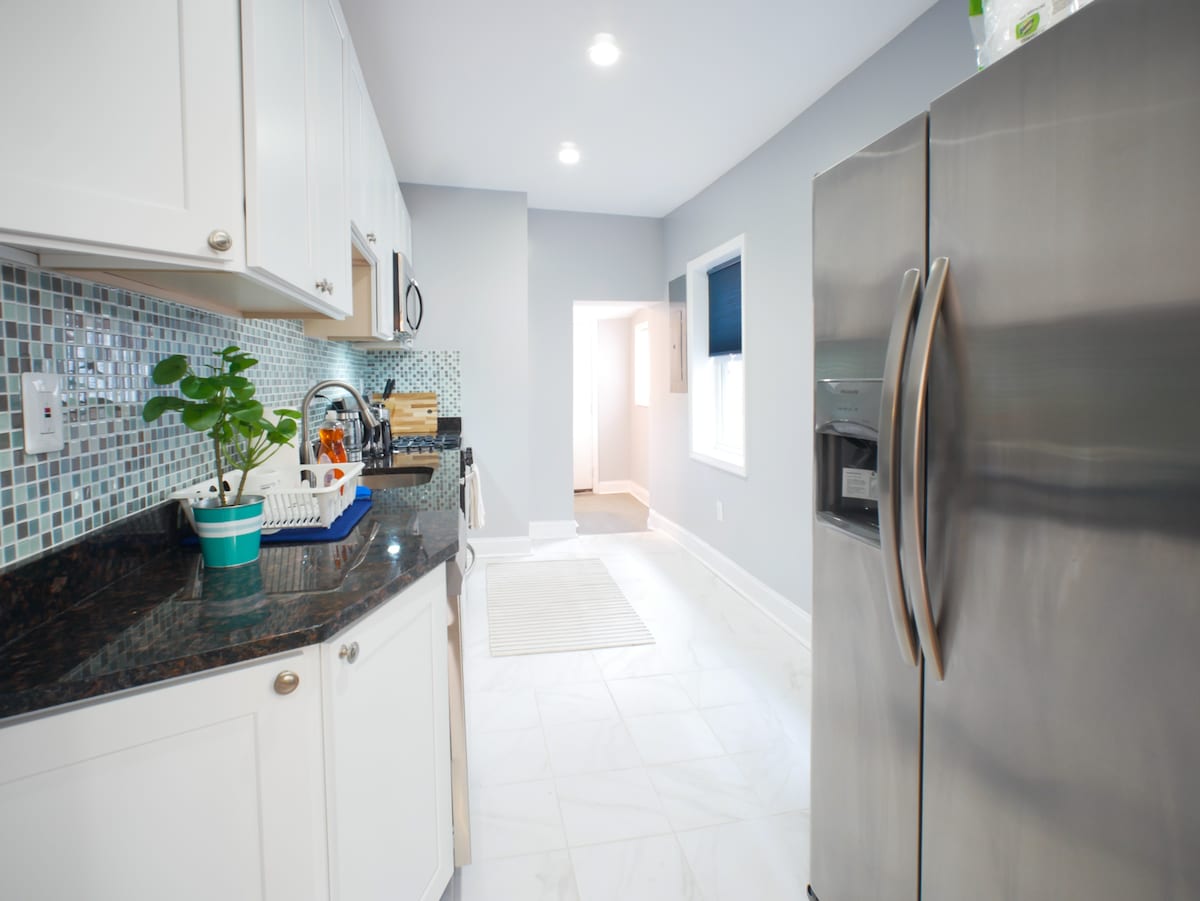 The kitchen features modern cabinetry and a sleek stainless steel refrigerator. A composed arrangement of dark granite countertops and a colorful mosaic tile backsplash adds character. Ample lighting illuminates the space, while a window at the end of the room offers natural light.