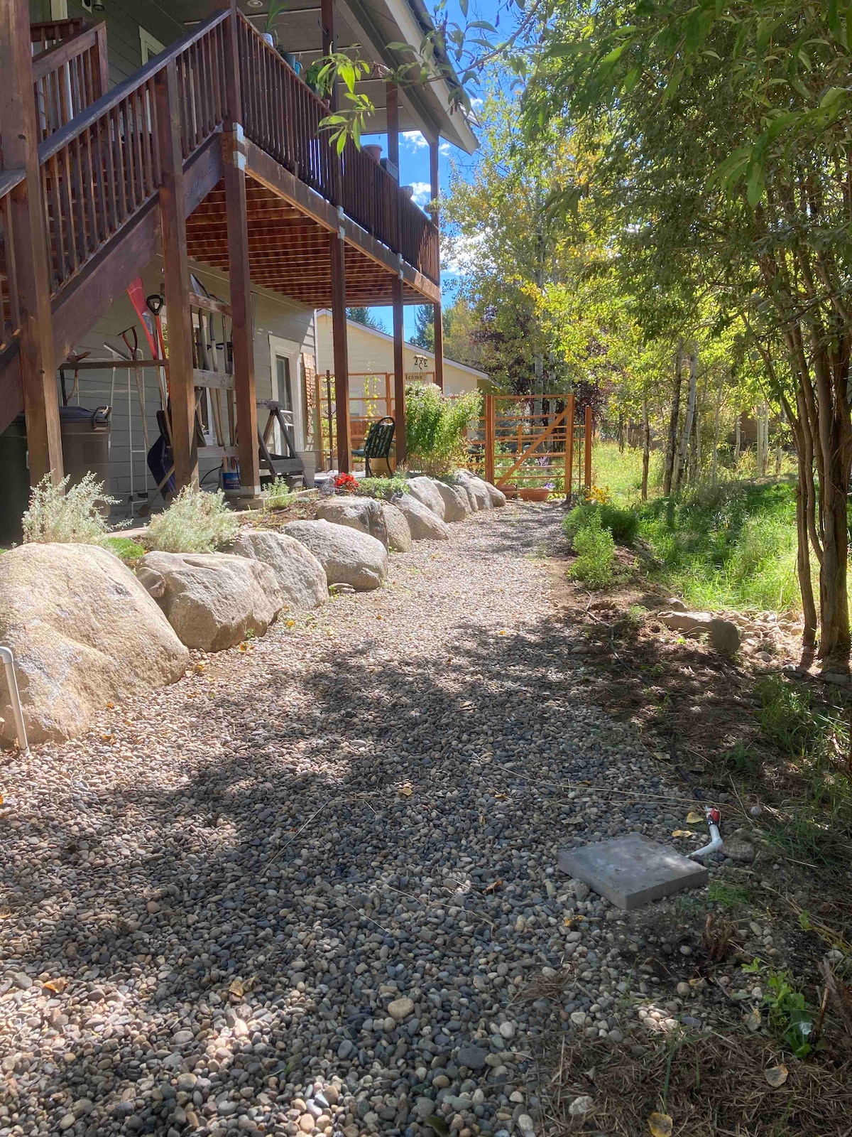 A gravel pathway runs alongside a landscaped area lined with smooth stones, leading towards a spacious deck and a lush green backdrop. The pathway is bordered by greenery and a wooden fence, providing a serene outdoor environment.