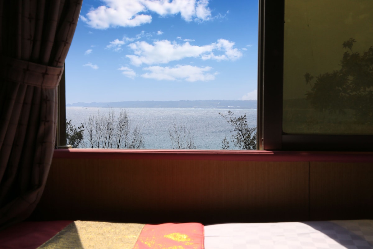 A large window frames a serene view of the ocean and sky, with soft clouds scattered above. The scene offers glimpses of trees along the shoreline, creating a peaceful atmosphere. The window is adorned with curtains that allow soft light to enter the room.