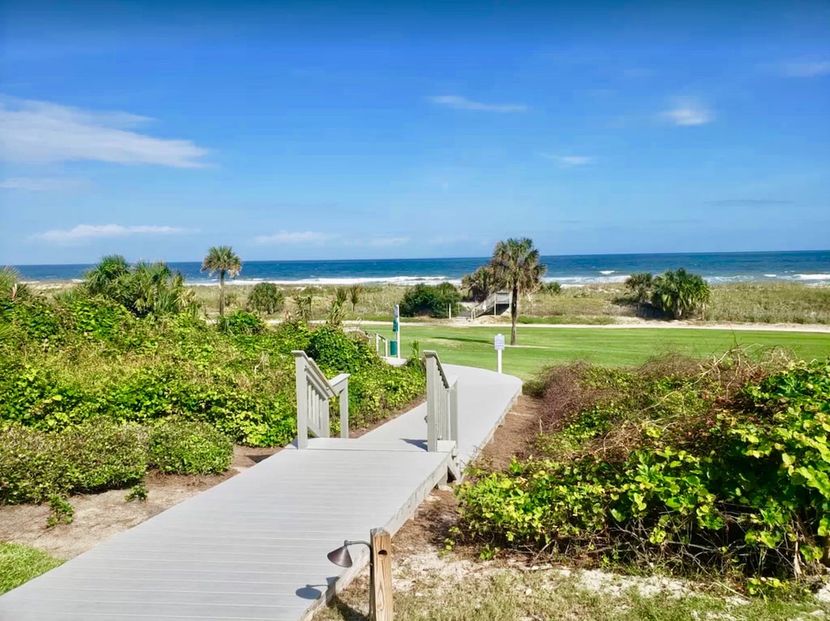 A wooden walkway leads towards the beach, bordered by lush greenery. The ocean is visible in the background under a clear blue sky, with beach grasses swaying gently in the foreground. Palm trees frame the scene, contributing to the coastal atmosphere.
