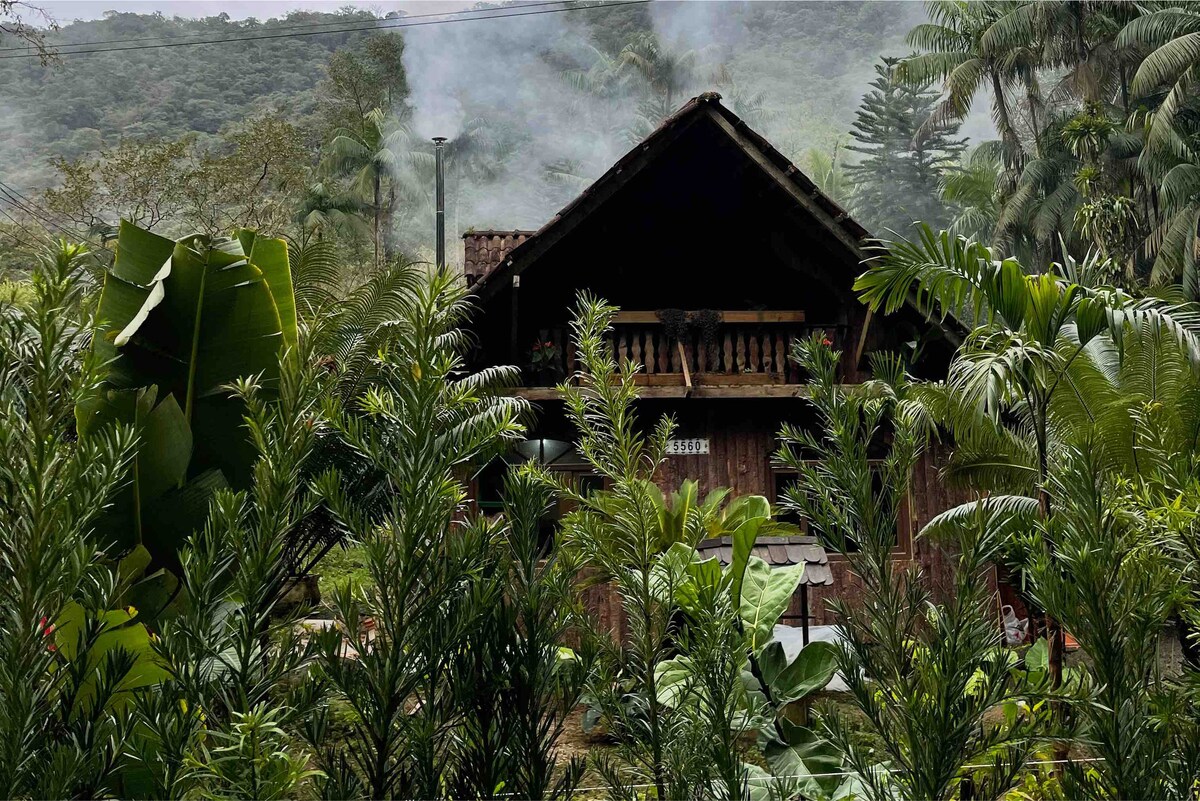 A rustic wooden cabin is partially visible behind lush green foliage, creating a serene and secluded atmosphere. Smoke gently rises from a chimney, suggesting warmth inside. The surrounding trees and plants emphasize the natural setting and tranquility of the environment.