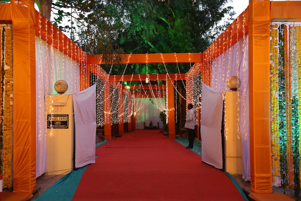 A vibrant walkway is adorned with orange drapery and twinkling lights, creating an inviting atmosphere. The path is lined with decorative pillars, leading towards a beautifully lit area. Lush greenery is visible in the background, enhancing the festive setting.