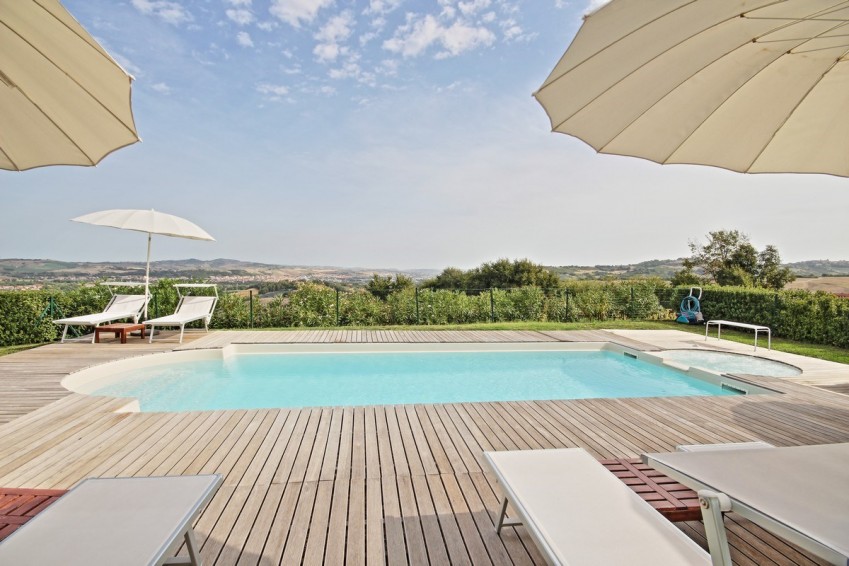 A serene outdoor space features a swimming pool surrounded by wooden decking. Lounge chairs are positioned for relaxation, and large sun umbrellas offer shade. Lush greenery and distant hills are visible, creating a calming backdrop under a clear sky.