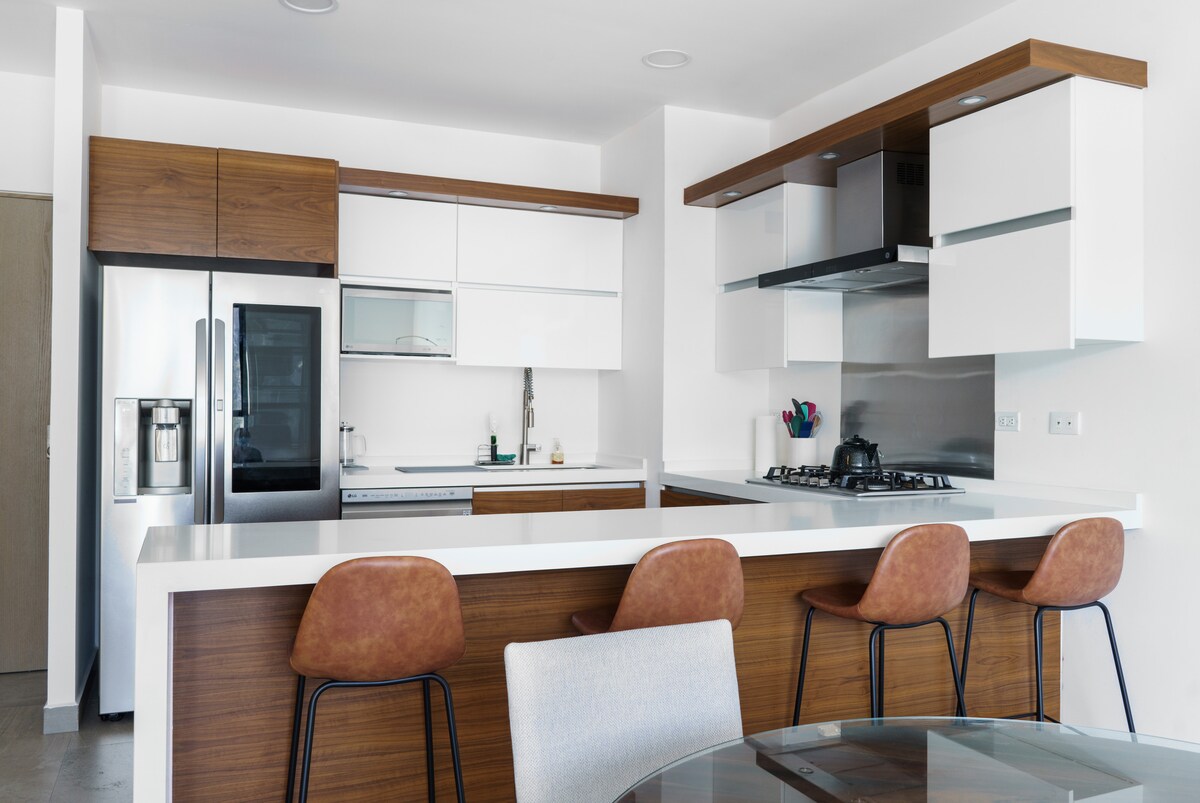 A modern kitchen features sleek cabinetry in light and dark woods, complemented by stainless steel appliances. A breakfast bar with several brown stools offers casual seating. The kitchen island includes a stove and ample counter space, providing a functional layout for cooking and dining.