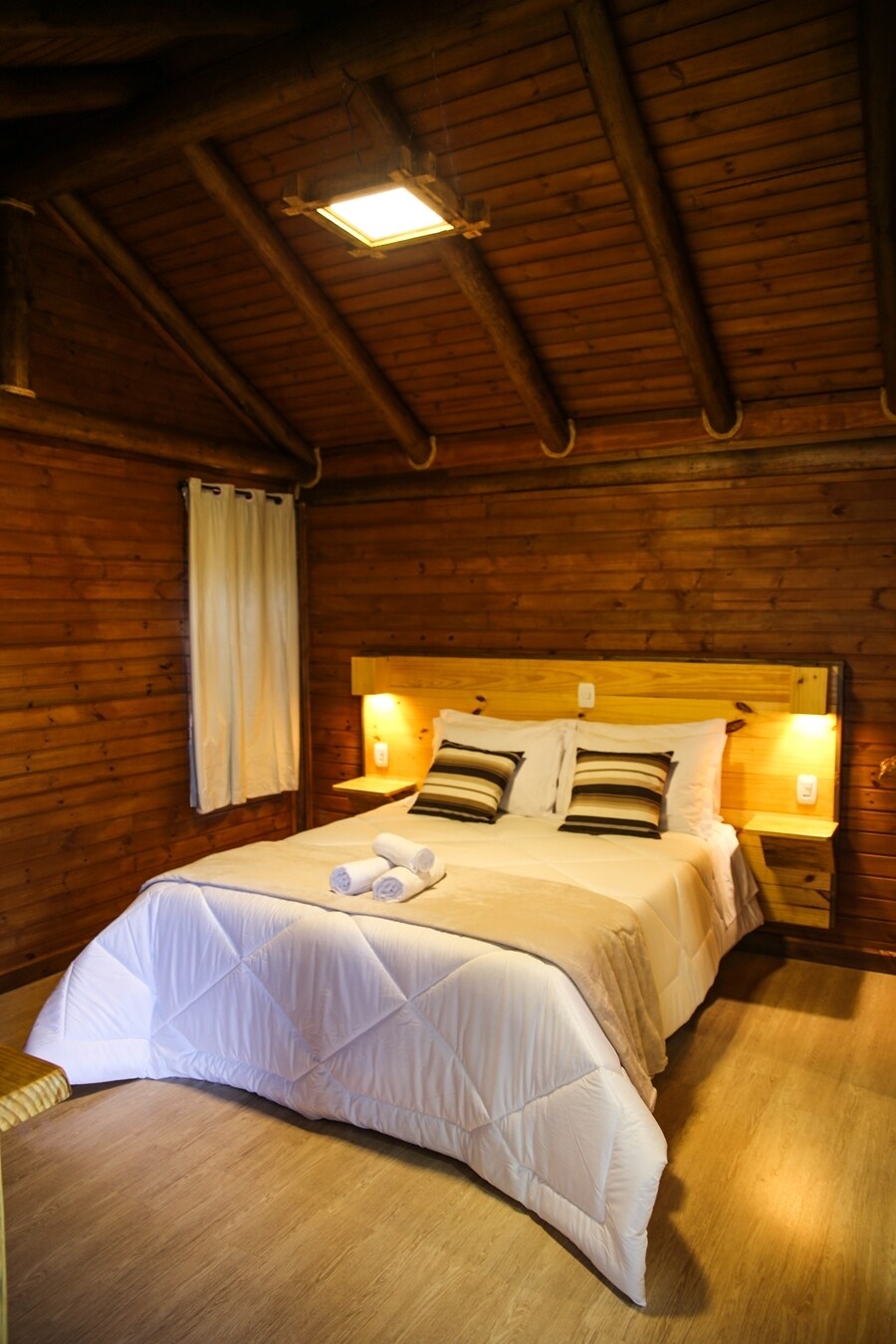 A cozy room features wooden walls and a vaulted ceiling, complemented by soft lighting. A spacious bed is dressed in white linens, with decorative pillows adding subtle contrast. A small towel arrangement is placed on the bed, and a chair sits in the corner.
