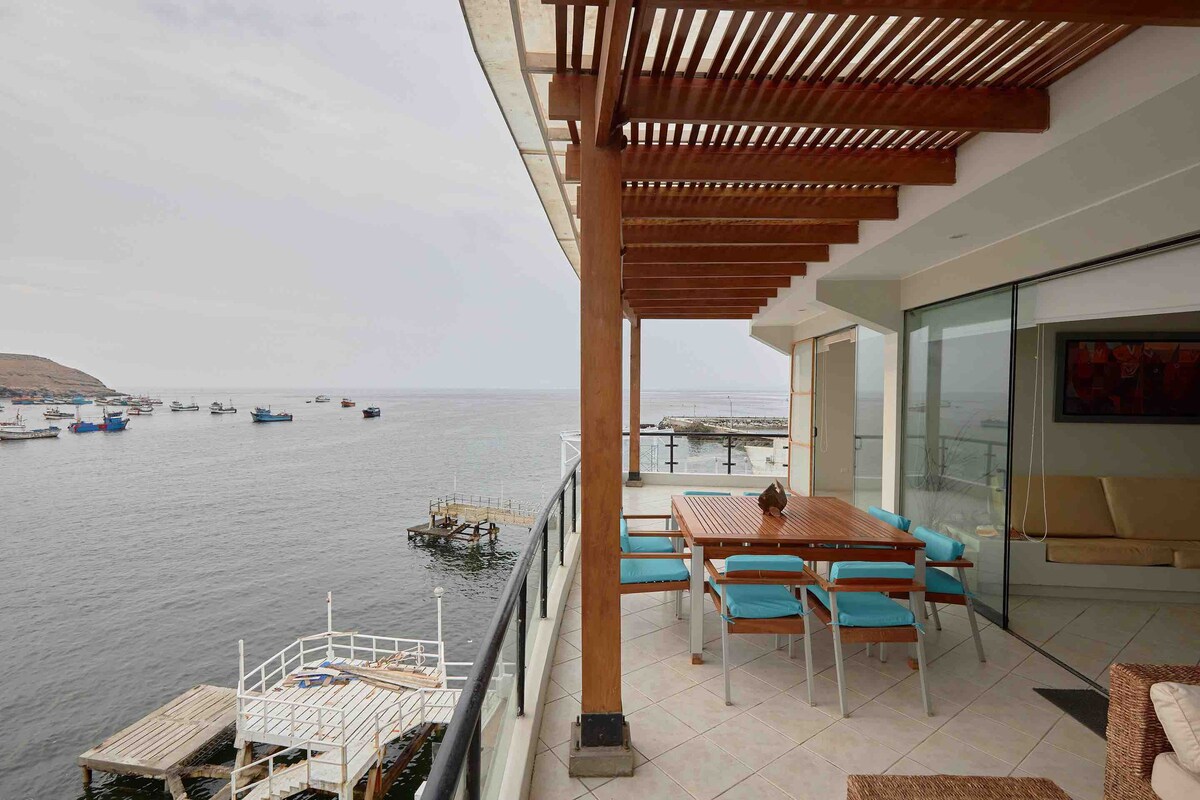 An outdoor terrace features a dining table accompanied by chairs, overlooking the serene waters. The structure is supported by wooden beams, providing shade. Boats can be seen in the distance, enhancing the coastal view while glass doors lead into the interior space.