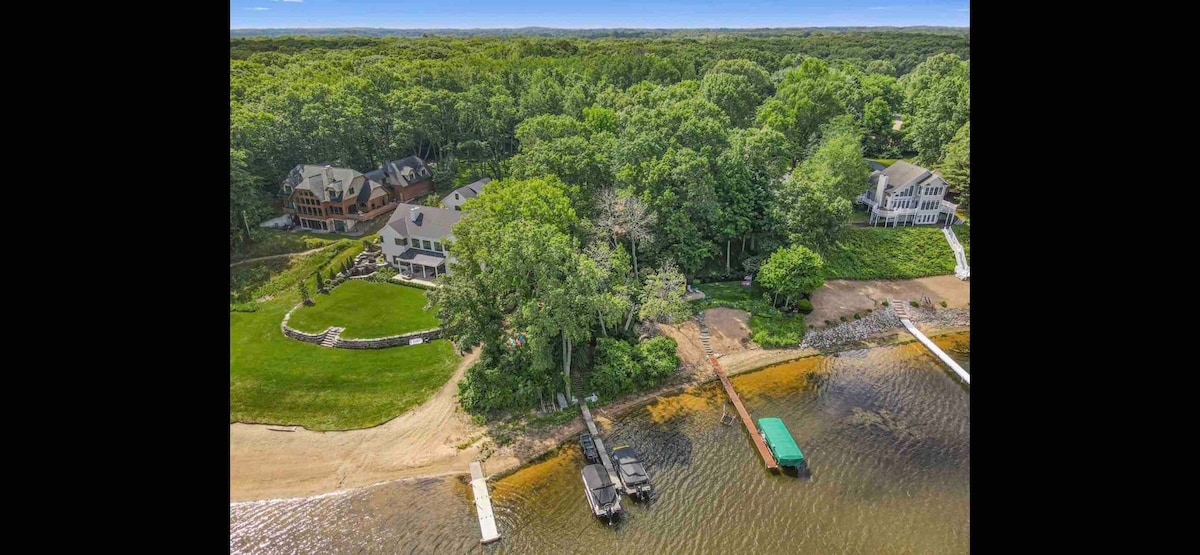 An aerial view showcases Red Oak Cottage nestled within a wooded landscape near the lake. The property features a sandy beach area, two docks, and a grassy lawn. The surrounding trees provide a sense of privacy, enhancing the tranquil setting.