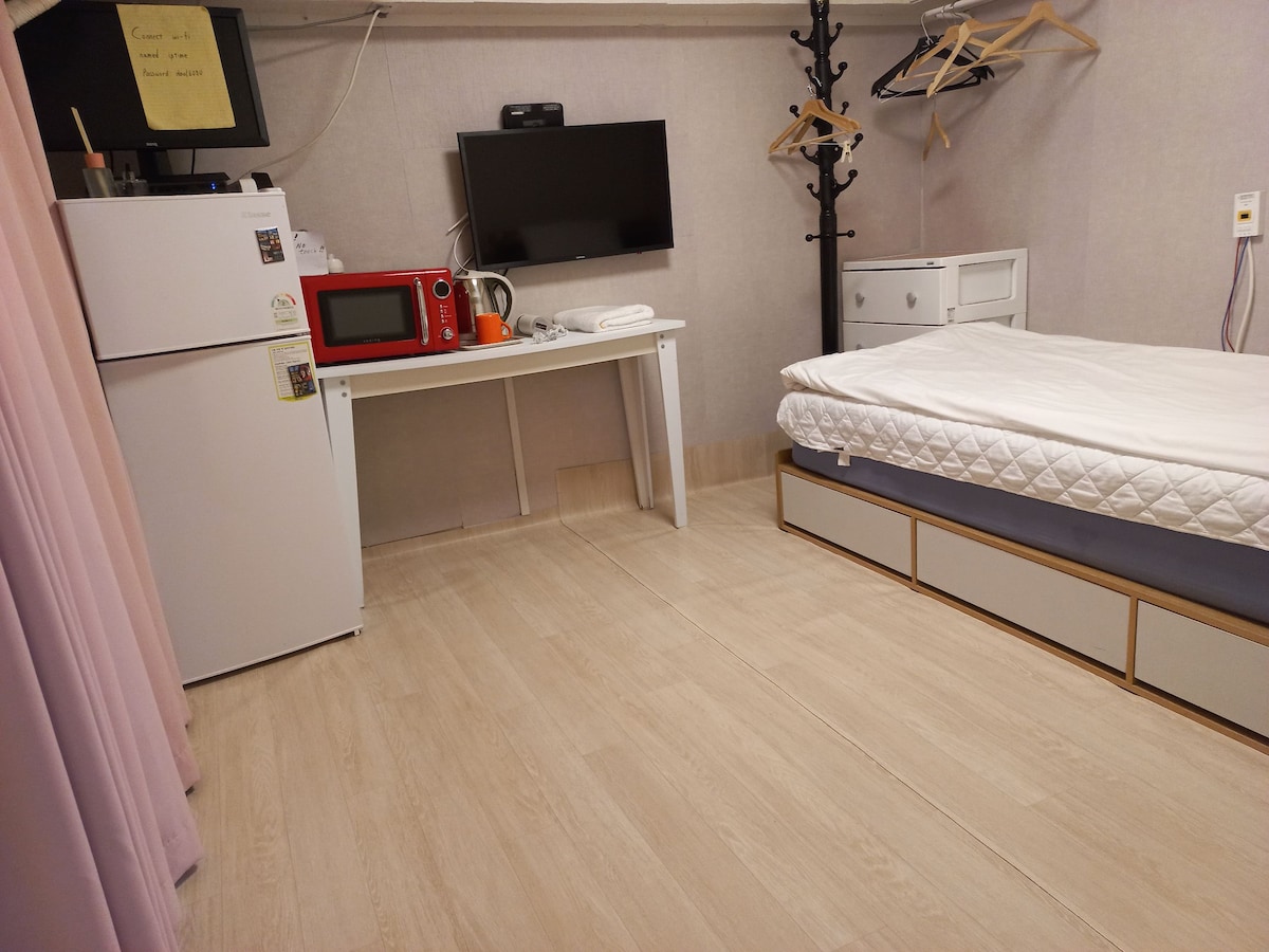A functional guest room features a spacious bed against a wall, complemented by light-colored flooring. A compact refrigerator and microwave are positioned on a small table, while a TV is mounted above. Additional storage is provided by a wardrobe and a hanging rack.